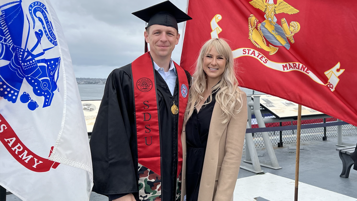 SDSU graduate Christopher Phillips in his cap and gown poses with his wife standing on the USS Midway, a historical naval aircraft carrier turned into a museum