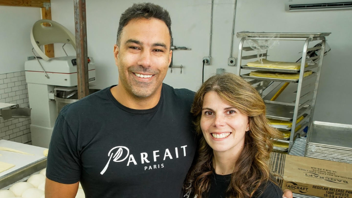 A man and woman wearing black t-shirts with Parfait Paris printed across the front are standing with their arms around each other with a sheet of dough and other baking implements behind them.