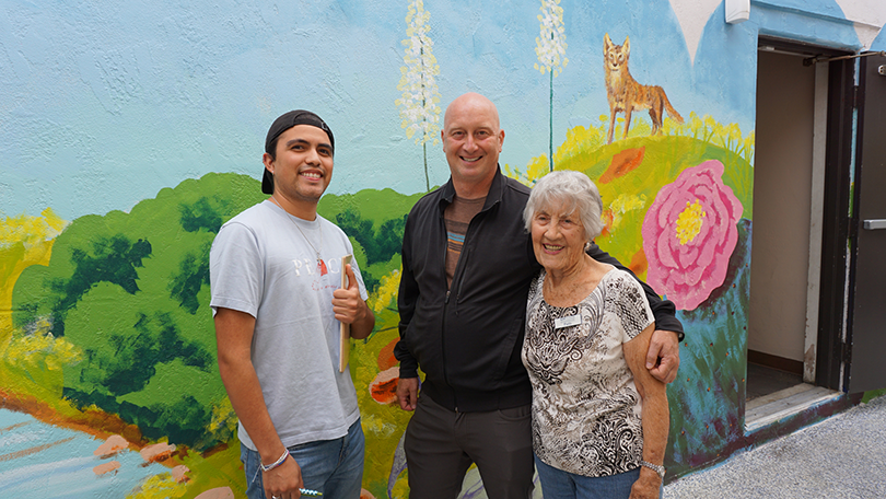 Two men and a senior woman are standing in front of a colorful mural depicting a green hill, giant flower, a coyote and blue sky.