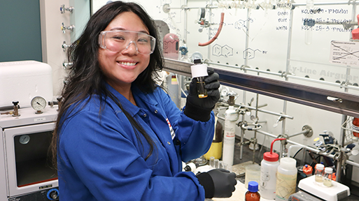 A young woman with shoulder-length black hair is wearing a blue lab coat and safety goggles in a lab full of equipment and whieboards. She is beamning as she holds up a small glass container.