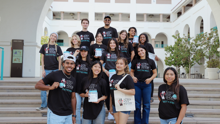 Fifteen young men and women, all wearing black t-shirts marked Aztecs Rock Hunger, stand in five rows of an outdoor staircase with a building in the background.