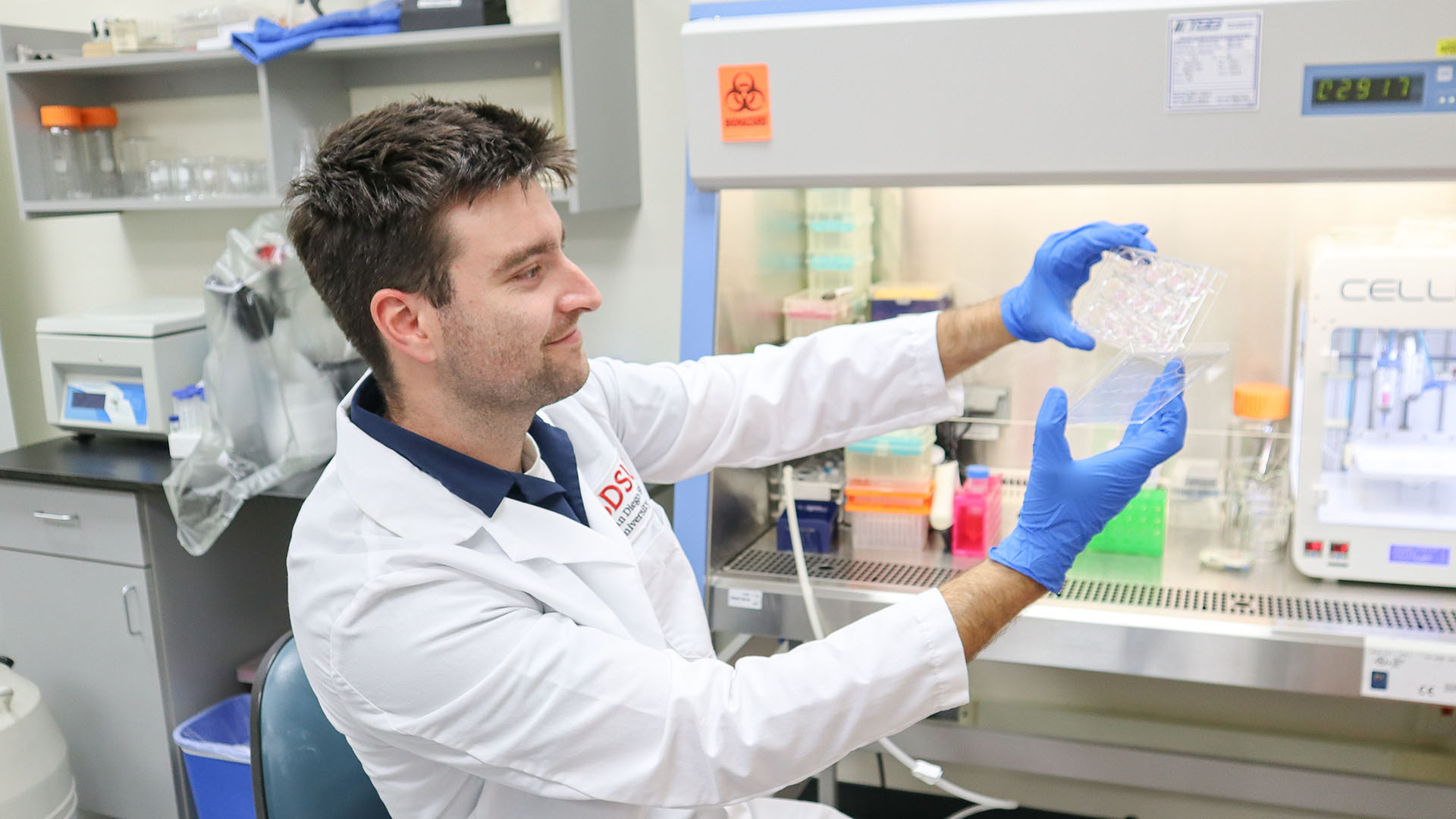 A young man seated in a laboratory and wearing a white lab coat and blue latex gloves holds a transparent lab instrument at arm's length.
