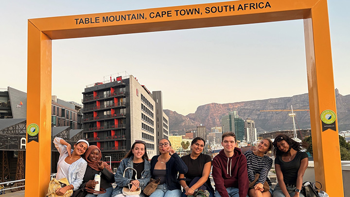 Eight young men and women are sitting on the bottom of an immense open wooden frame labeled at the top as Table Mountain, Cape Twon, South Africa, seen as the background.