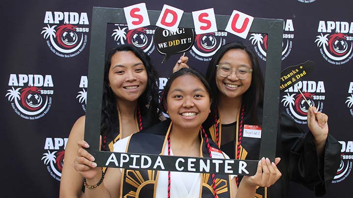 Three young Asian women pose for a photograph inside a frame, held by the woman in the center, that reads SDSU APIDA CENTER. Two of them word balloons that read, OMG! We did it! and Thanks Mon & Dad.