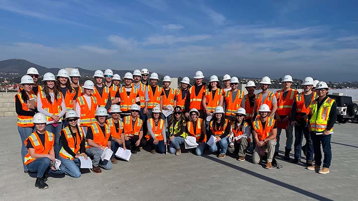 Three dozen people, mostly young, stand in two rows on a concrete site with the front row squatting. All are wearing white construction hardhats and reflective safety vests. In the background is a blue sky with light clouds and some distant hills.