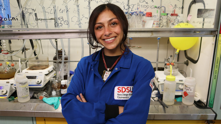 SDSU senior Alyssa Gomez is photographed in a blue lab coat, smiling with arms crossed, in her biochemistry laboratory.