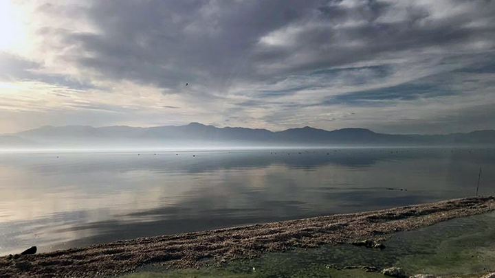 A vast, calm lake is seen from a rocky shoreline. It reflects an overcast sky with a glimmer of sunlight to the left. In the distance light hovers over the surface, and a ridge of mountains.