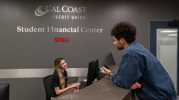 A young man in a deep-blue denim shirt or jacket is leaning across a counter toward a young woman seated at a reception station. Silver lettering on the wall behind her reads Cal Coast Credit Union Student Financial Center, with SDSU below it in red.