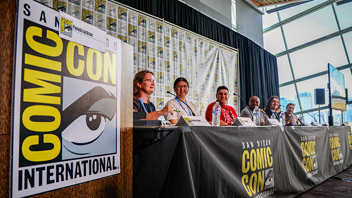 In hall with floor to ceiling windows overlooking downtown San Diego, SDSU faculty and staff sit at a table to take part in a panel during Comic-Con 2023.