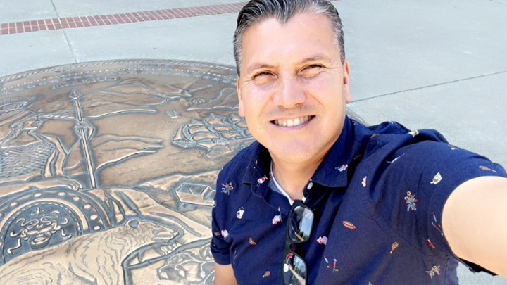 Francisco De La Rosa wearing a blue woven shirt takes a selfie outdoors in front of 