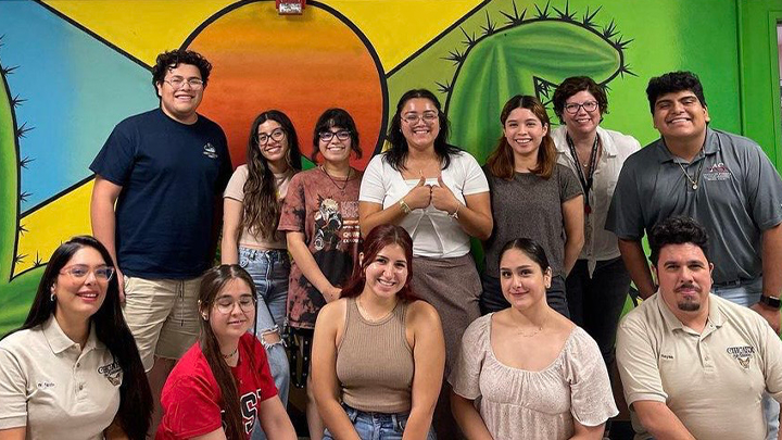 Twelve people, with one row standing and the other squatting in front of them, pose in front of an indoor mural. In yellow, blue, orange and green it depicts two cacti and an orange sun.