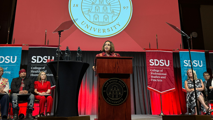 The president of San Diego State University stands at a podium on a stage with the bottom half of the university logo on a screen behind her, and two banners representing individual colleges on either side.