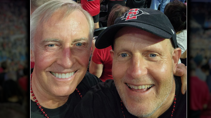 Two men with their heads close together pose for a photo. One is wearing a black SDSU interlock baseball cap.