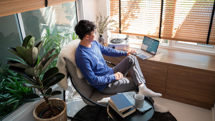 A young man with dark hair, wearing a blue sweater and tan plants, sits at a corner desk in front of a tall window and performs work on his computer.