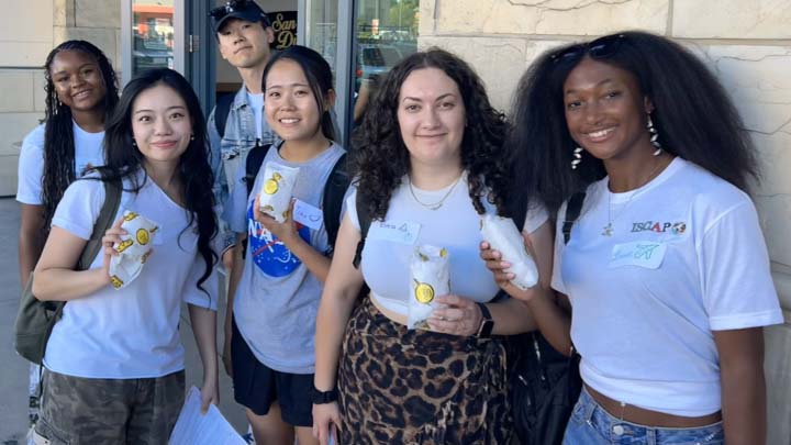 A young, multiracial group of one male and five female students are standing along the outside wall of a store. Four of them are holding food items in takeout pouches.