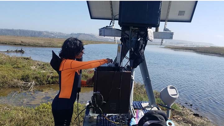 A young person, seen in profile with black hair obscuring most of their face, is working on a laptop keyboard placed on top of a large instrument, about four feet high. Behind them we see an estuary, forking off into two branches, surrounbded by flat wetlands.