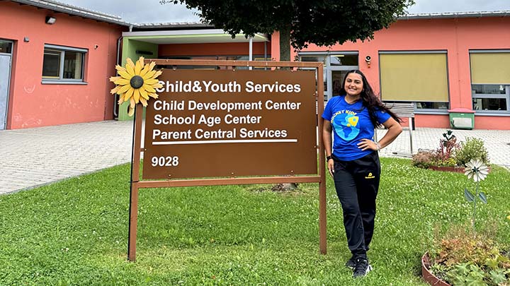 A young woman wearing a blue t-shirt with a Rocket Kids logo is standing on green grass beside a sign for Child and Youth Services, with an image of a sunflower attached at upper left. A one-story building with large windows is behind her. 