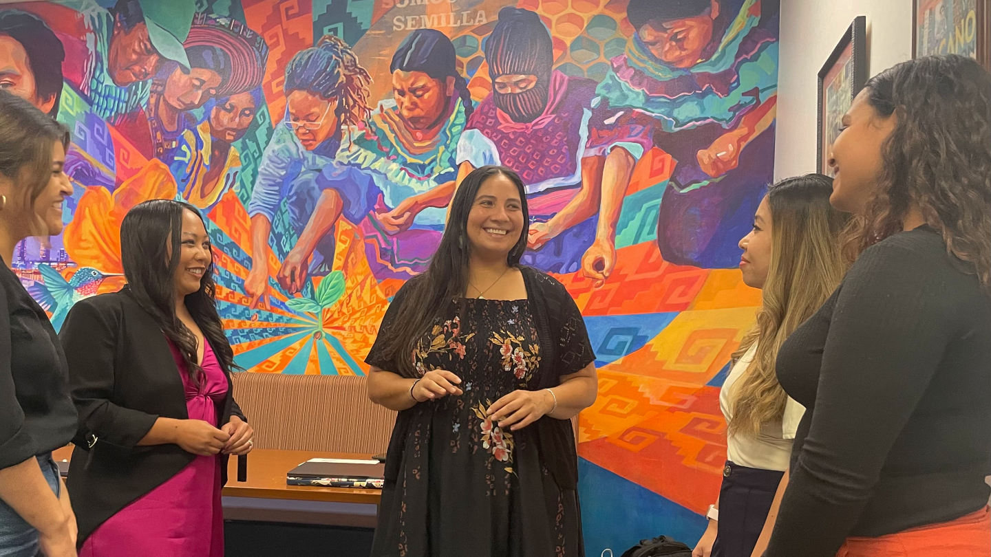 A woman in a black floral print dress, with black hair below her shoulders, is smiling and standing between four other women in front of a colorful mural of Indigenous farmworkers.