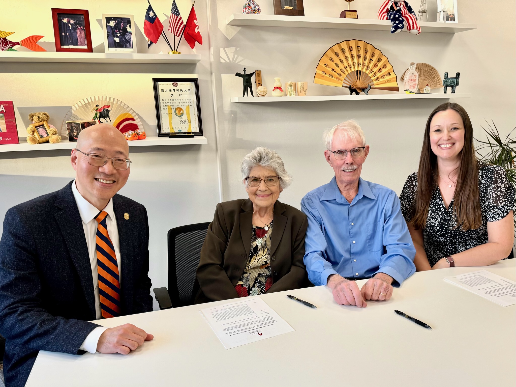 Four people are seated at a table with pens and a document in front of the middle two, a senior couple. A man in a business suit is on the left and a woman in a white and black patterned dress is on the right. Behind them are shelves of framed photos and knickknacks.