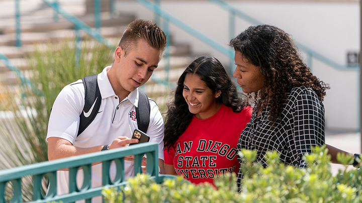 Three students are seen conversing as a male on the left shows the screen of his smartphone to two women, one of whom is wearing a red San Diego State University sweatshirt. They are standing along a teal railing with light landscaping, and addiitonal landscaping and a blurry staircase behind them.