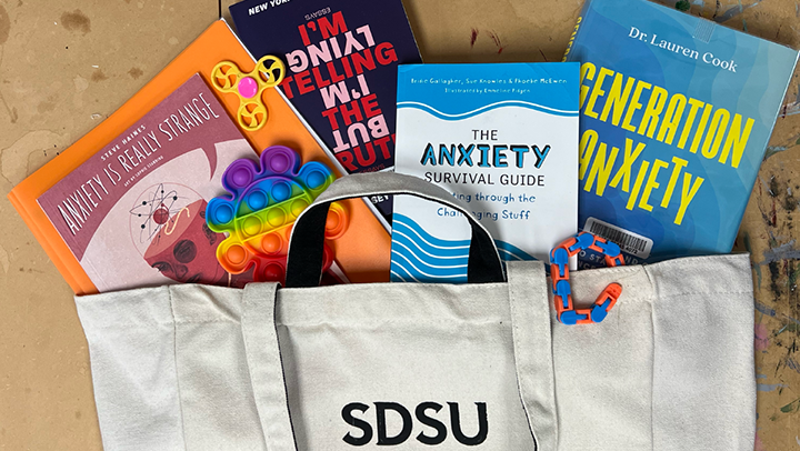 An image of SDSU's Pick Me Up Kit for SDSU students shows a bag filled with books about anxiety and puzzles. 