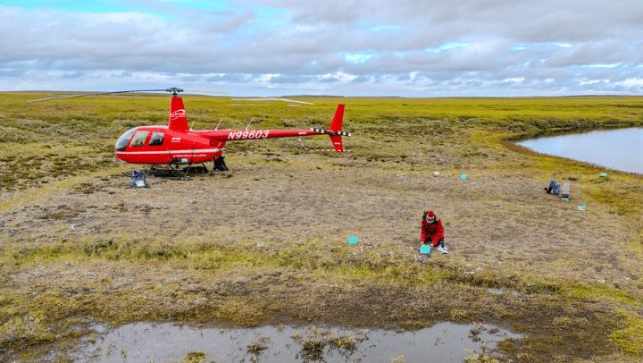 A photo of a red helicopter and researcher in an empty field taking a sample of soil.