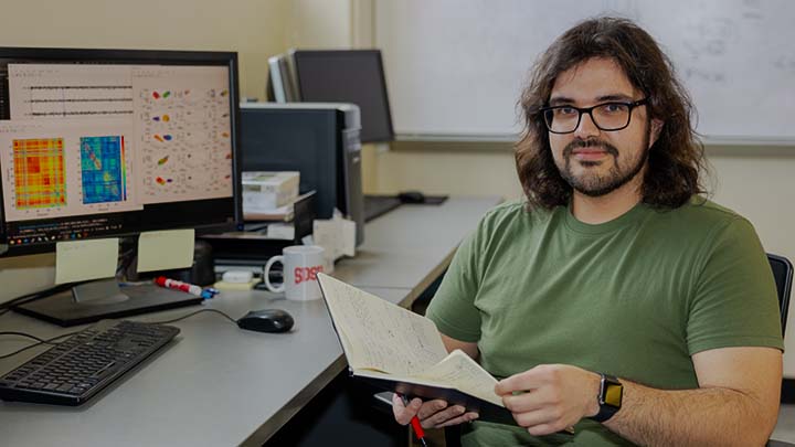 A young man with shoulder-length brown hair and a light beard is sitting at a table with a computer model and a keyboard, holding a notebook in both hands and a red pen in his right hand. He is wearing a plain green short-sleeve t-shirt and is looking directly into the camera.