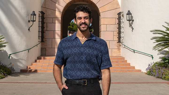 A young man stands on the pavement outside a building, San Diego State's Hepner Hall, framed precisely in the middle of its arched entryway. He is wearing a blue short-sleeve shirt and has his right hand tucked inside a pocket, his thumb on the outside.