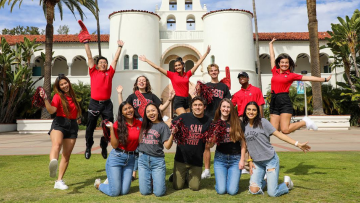 Twelve young men and women, all dressed in San Diego State gear, are posed in two rows with Hepner Hall in the background. Three of them are in midair, jumping with arms outstretched.