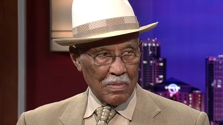 A senior man is seated in a televison studio, wearing a jaunty fedora and a grey suit with a necktie. He is looking at an off-camera interviewer, with a skyline backdrop,