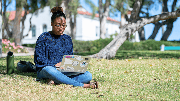 A student sits on a rolling hill using a computer and trees in front of large white stucco spanish-style buildings fill the background