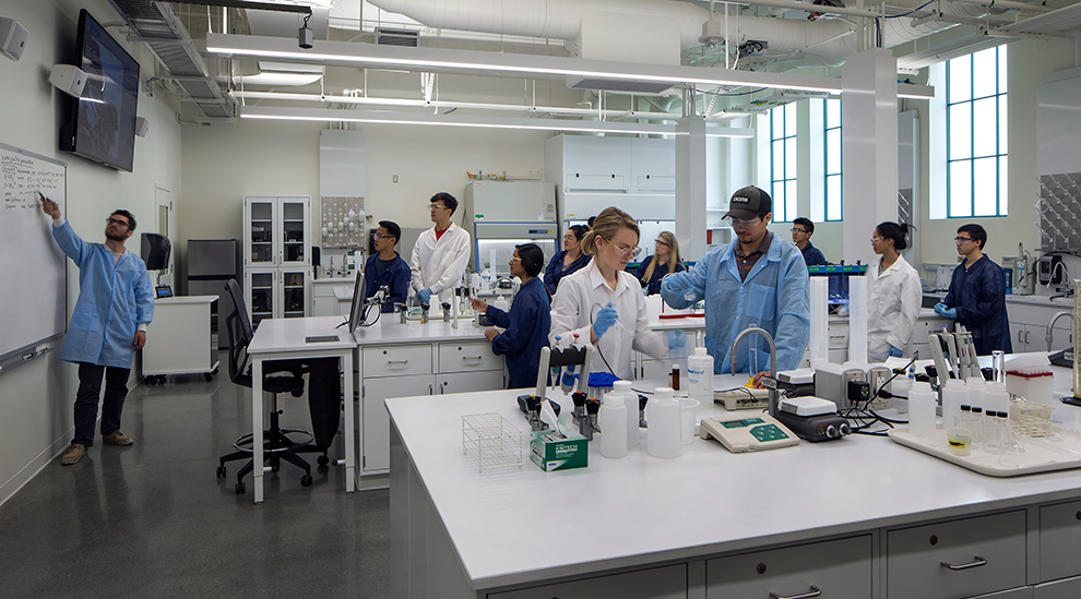 In a university lab 10 students wearing lab coats stand at two long tables as an instructor gestures toward writing on a whiteboard.