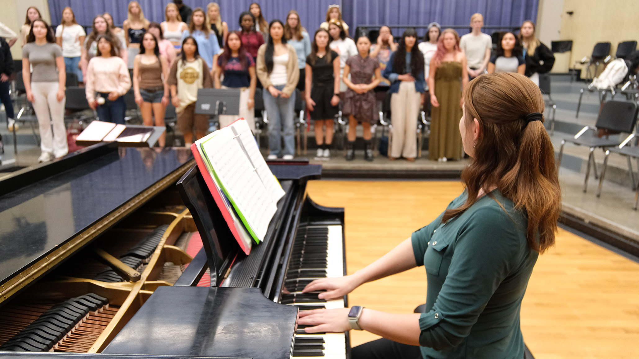 A woman dressed in green with her head turned away sits at a piano, both hands on the keyboard, and looks across the room to a group of 30 or more female students on risers, in three rows.