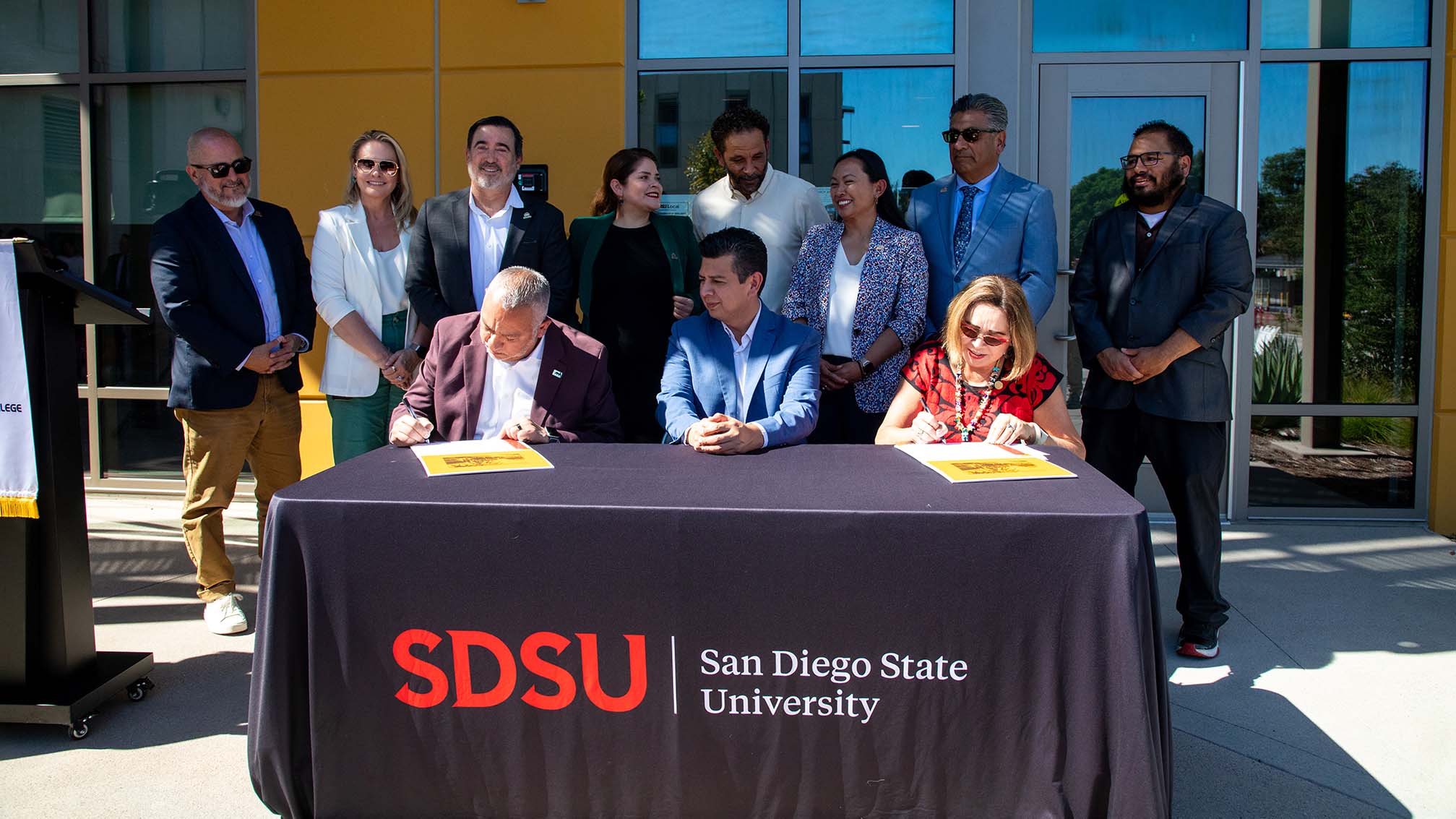 Three people are seated at a table with a banner reading SDSU San Diego State University. A man and woman on either end are signing documents while the man in the middle looks on.