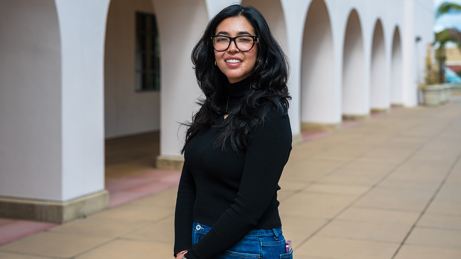 A young woman with black hair descending below her shoulders, wearing a black top and blue jeans, stands with her left shoulder facing the camera in front of a building with arches at street level.