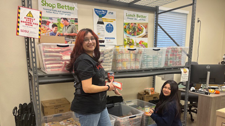 Two women, one standing and one squatting, turn toward the camera in front of shelves with food bins and posters that read Shop Better and Lunch Better.