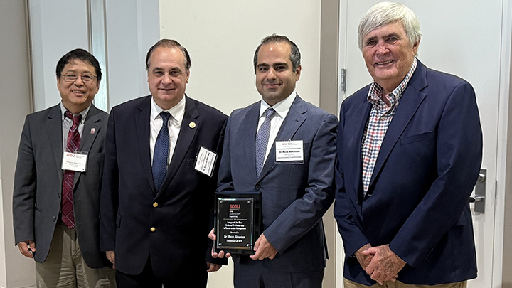 Four men in business suits stand shoulder to shoulder. The third from the left is holding a small plaque.