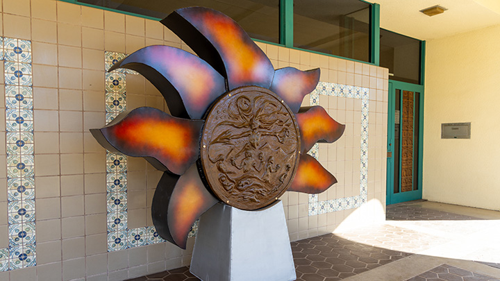 A large sculpture on a pedestal with seven red-orange rays emitting from a circular center that depicts people and elements of an Indigenous culture.