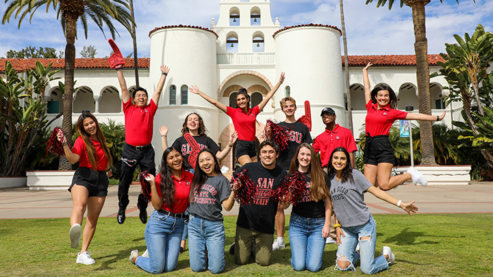 Twelve young men and women on a green lawn pose in two rows, all wearing either red and black clothing or tops with San Diego State University logos. A Mission Revival style building is behind them.