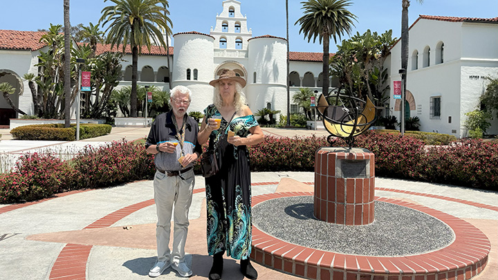 A man and a woman pose on a platform with an astronomical instrument on a platform and a Mission Revival-style building in the far background.