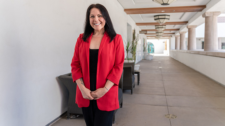 A smiling woman wearing a red blazer over black clothing stands in the outdoor corridor of a building with columns between a wall and ceiling, wooden beams and lighting fixtures.