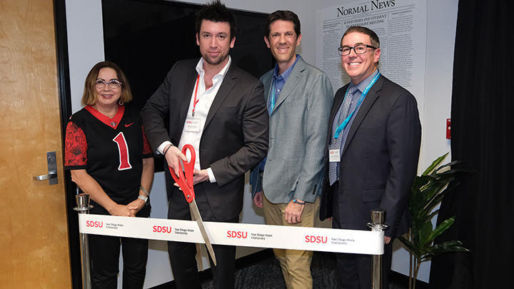 A woman and three men lined up behind a ribbon between two stanchions. One of the men is holding an oversized pair of scissors, about to cut the ribbon.
