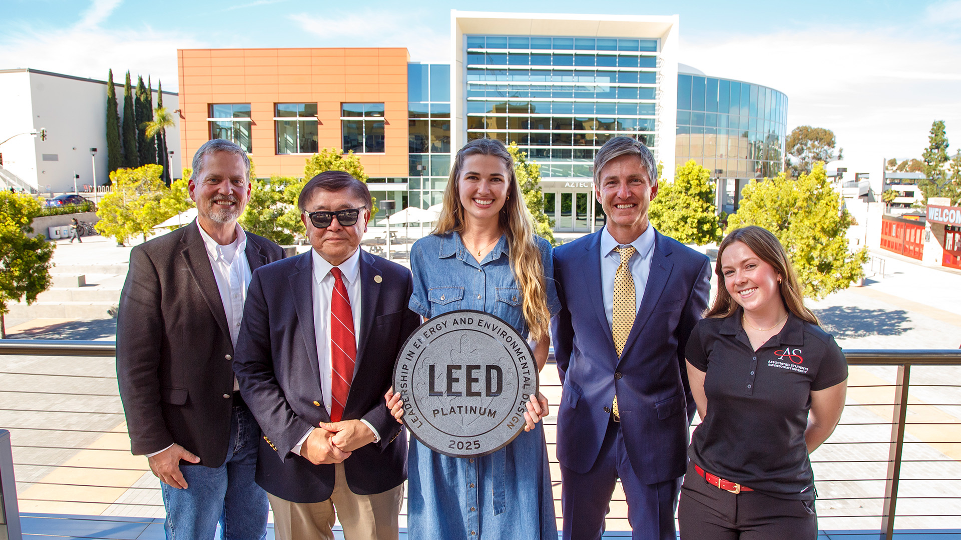 Three men and two women stand on a second-story terrace overlooking a campus. The woman in the middle holds a placard reading LEED Platinum 2025.