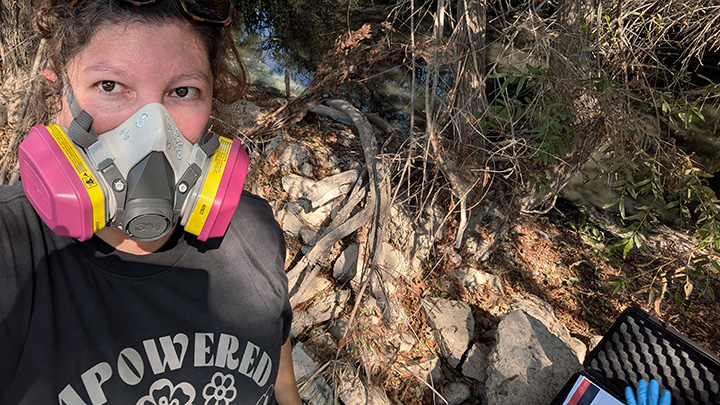A woman wearing a half-face respirator with dual cartridge filters stands on a riverbank with thick vegetation behind her.