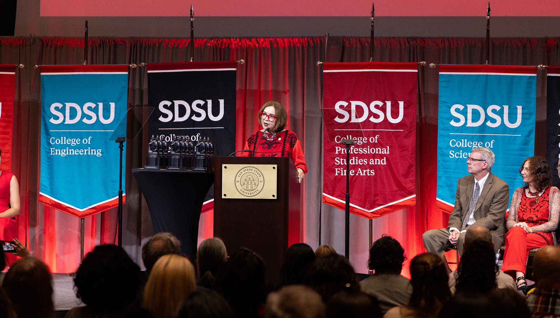 A woman in a red and black dress delivers remarks from a podium with a San Diego State University plaque on it and four banners behind her.